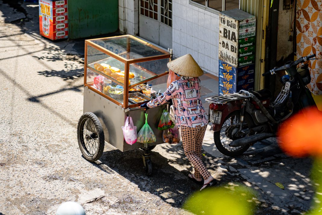A street vendor wearing patterned clothing and a conical hat pushes a small food cart with a glass display case filled with baguettes and prepared dishes