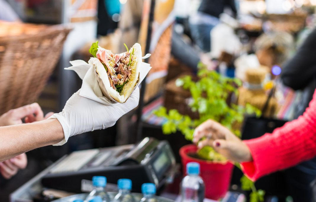 A vendor wearing a white glove hands a freshly prepared wrap filled with meat, lettuce, and cheese to a customer at Taste of Las Cruces, capturing the lively exchange of food and community spirit.
