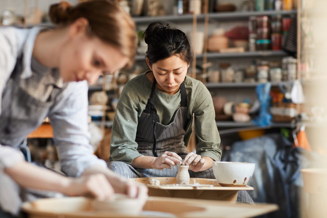 Two individuals work with clay on pottery wheels in a ceramics studio, shaping vessels with focus and care. Shelves of pottery pieces and tools surround them, highlighting the creative process of Pueblo-inspired ceramics.