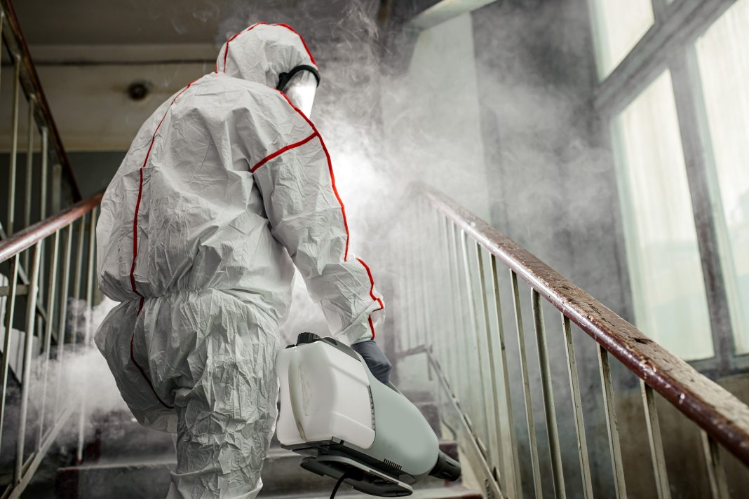 A healthcare worker wearing protective gear disinfects a rural cabin, spraying bleach on rodent droppings to prevent hantavirus transmission.