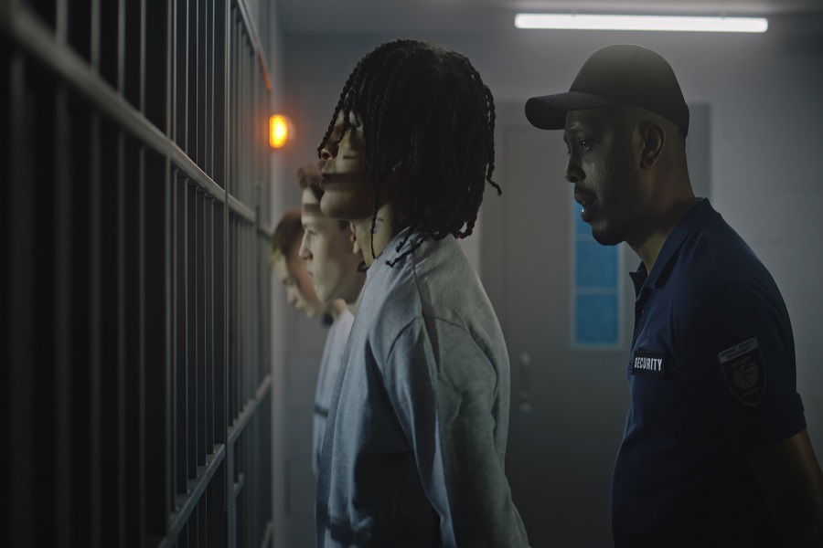 Group of teen prisoners stand in front of jail cells, warden talks with them. Multi ethnic teenagers serve imprisonment term for crime in detention center. Young criminals in prison. Justice system.