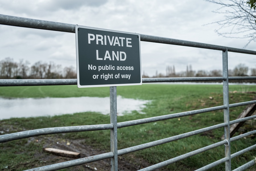 Close up of a newly installed Private Land metal sign at the gated entrance to farm pasture and a rural conservation area in the UK. The land is partially flooded due to persistent heavy rain.