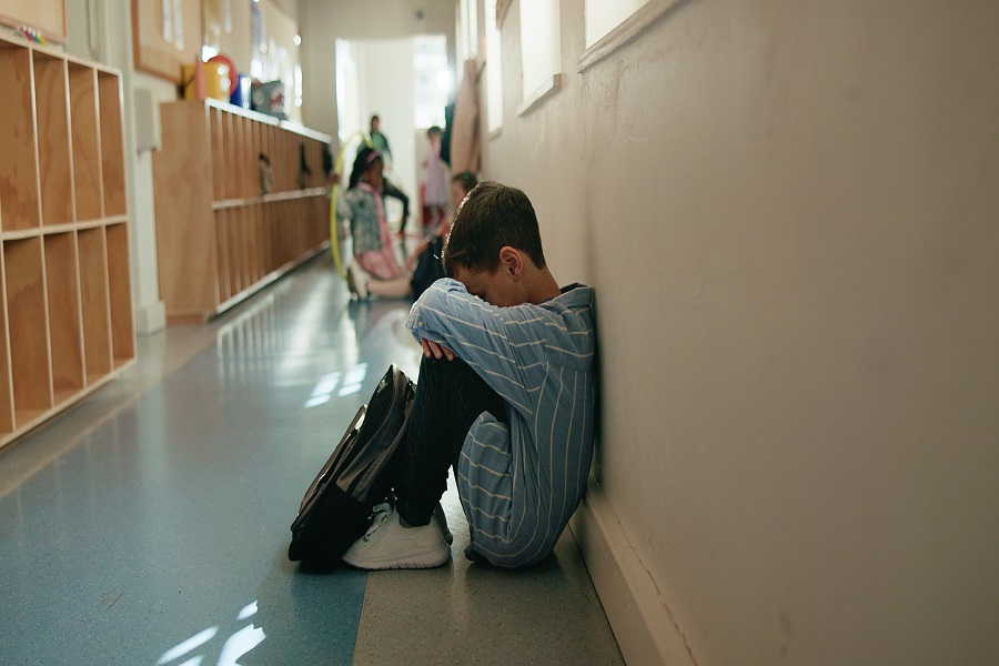 Boy, sad and depressed in corridor at school, alone and social isolation with mental health on floor. Child, anxiety and bullying in hallway with profile, frustrated or crying with stress at academy.