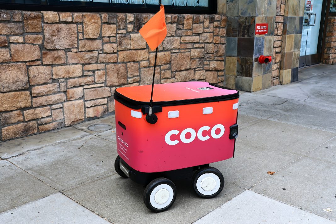 An autonomous DoorDash delivery robot travels along a sidewalk while transporting a food order in an urban area.