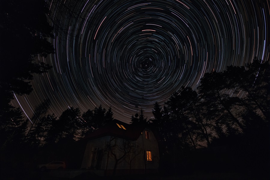 House surrounded by trees under dark night sky. Movement of stars around pole star on north hemisphere. Startrails on night sky, long exposure composition.