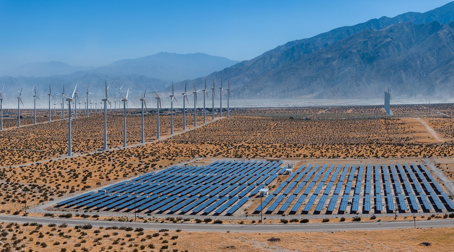 Renewable energy landscape with large wind turbines and solar panels in a sunny, arid desert valley, surrounded by mountains, under a clear blue sky.